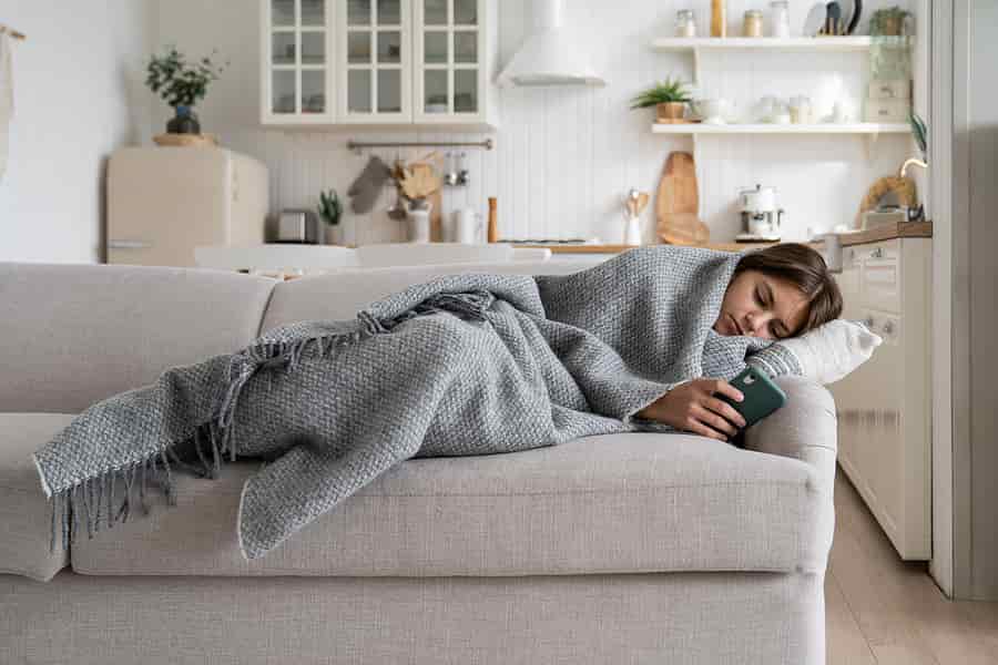 Depressed teen girl laying on the coach in her house wrapped up in a blanket and scrolling on her phone